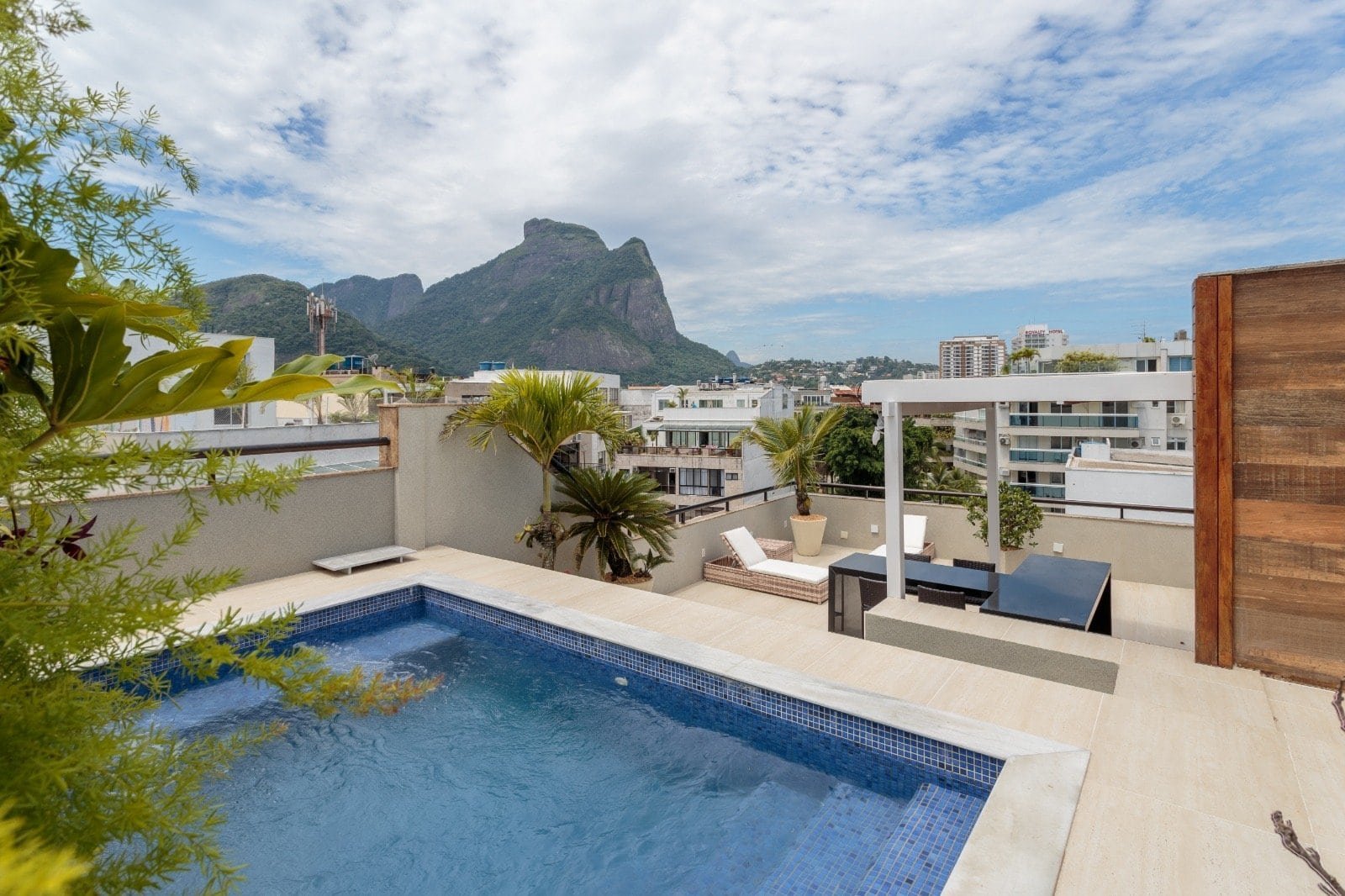 Piscina na varanda de imóvel com vista para montanha e skyline urbano, decorada com plantas tropicais e móveis de lazer, ideal para momentos de relaxamento e entretenimento ao ar livre.