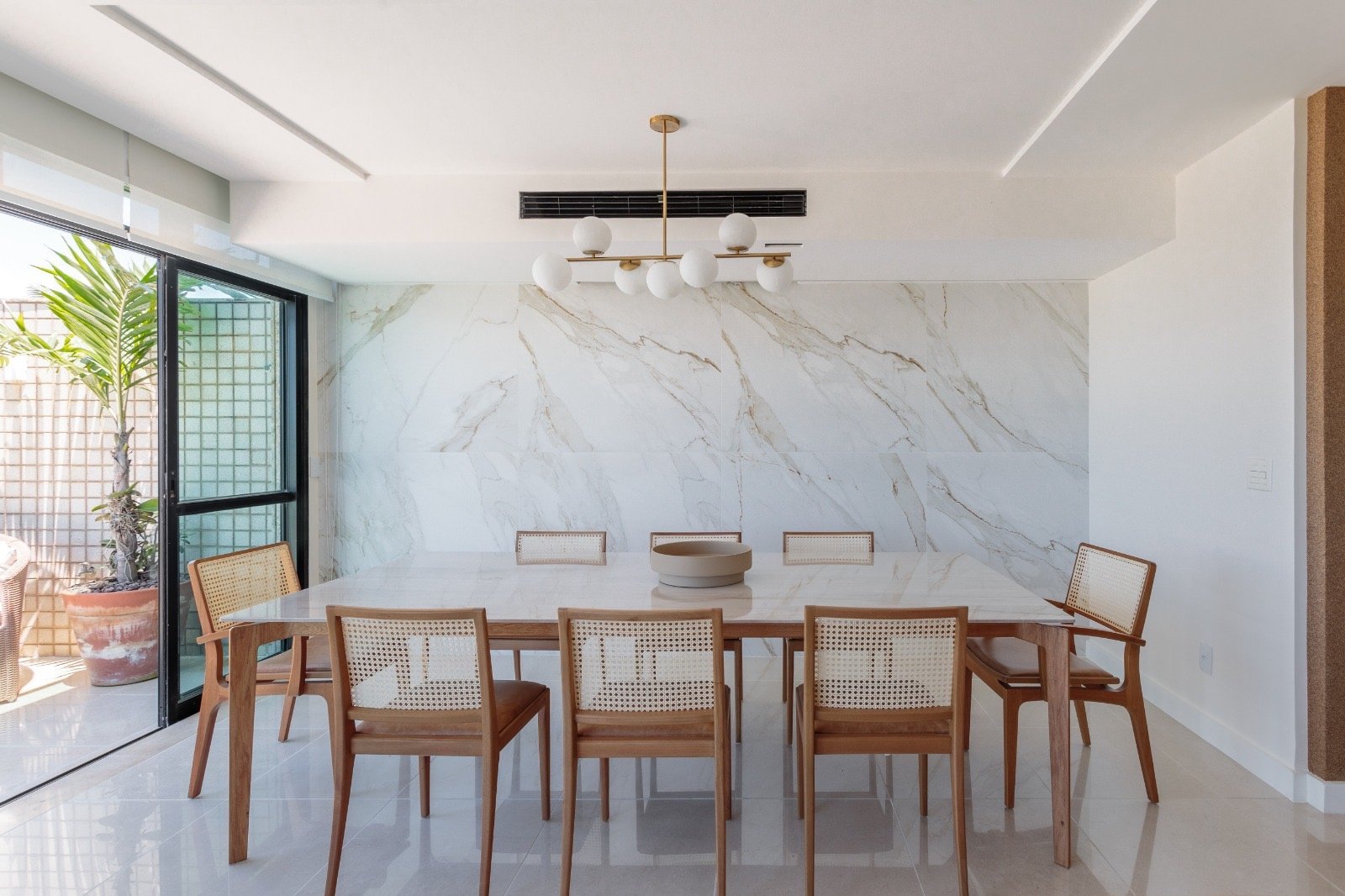 Moderno ambiente de sala de jantar com mesa de mármore, cadeiras de madeira com assento de rattan, iluminação moderna e parede de mármore clara, perfeito para eventos corporativos e reuniões elegantes.