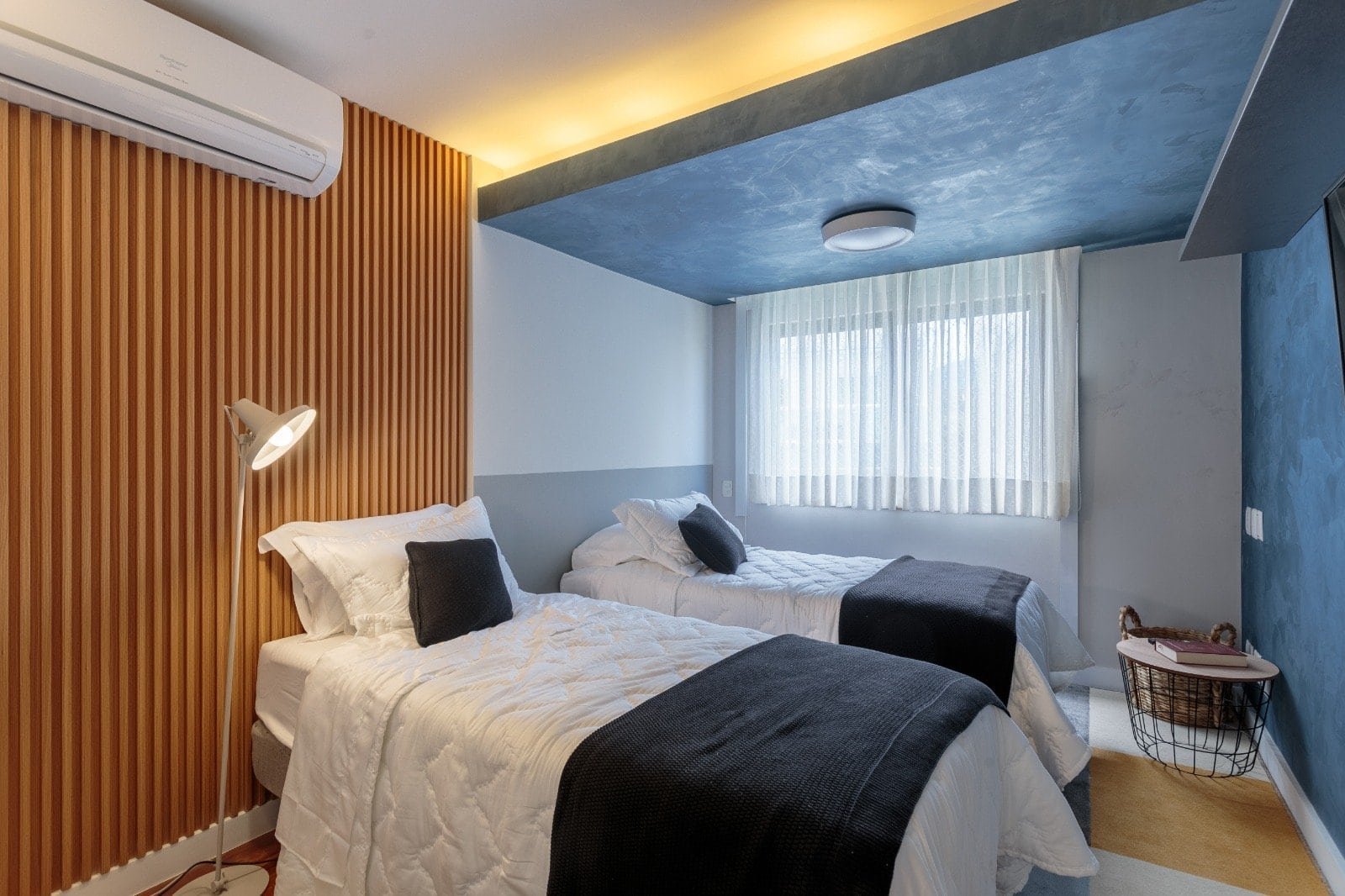 Conjunto de duas camas de solteiro com roupa de cama branca, almofadas pretas, parede de madeira na cabeceira, iluminação de leitura, janela com cortinas brancas, teto azul com textura e iluminação suave, decoração moderna e aconchegante.