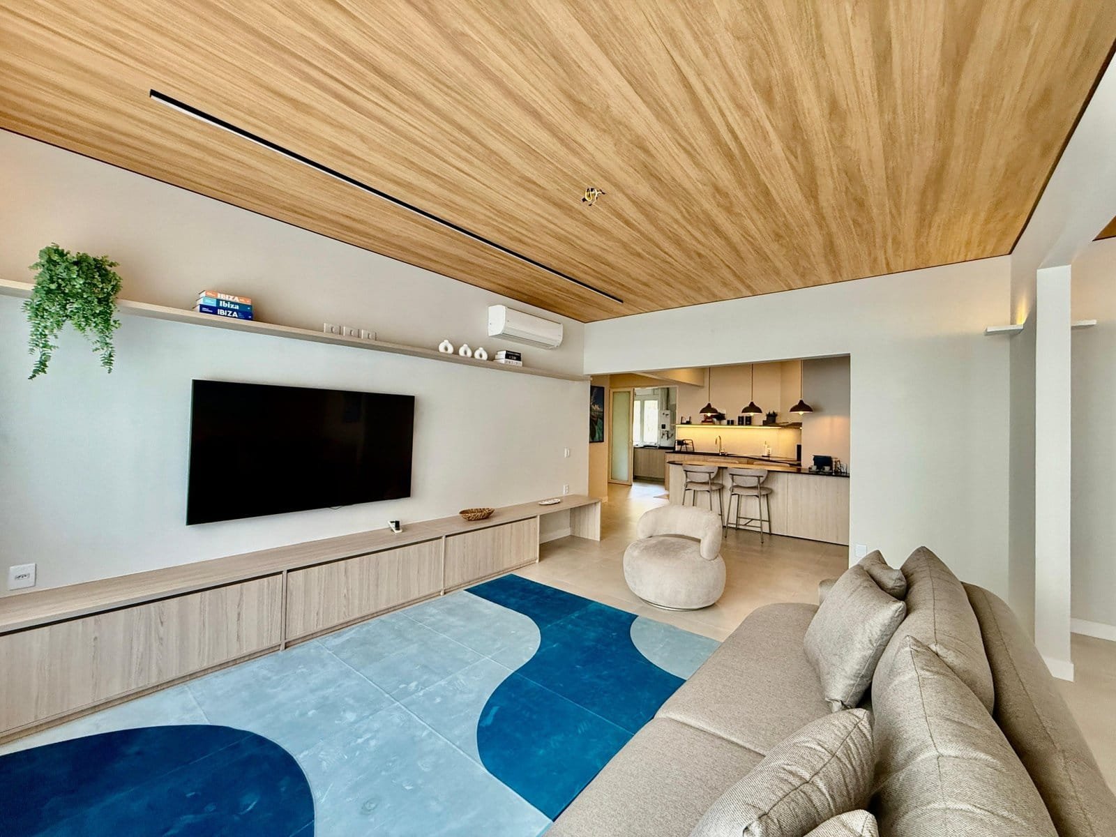 Moderno apartamento com conceito aberto, sala de estar com televisão, decoração minimalista, teto de madeira, espaço integrado com cozinha e uma área de convivência aconchegante.