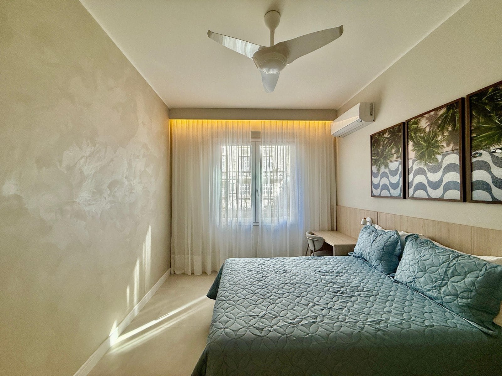 Conjunto de camas confortáveis com roupas de cama em tom azul, iluminação suave, decoração com quadros de palmeiras, janela com cortinas brancas e ar condicionado, ambiente de quarto de hotel moderno e acolhedor.