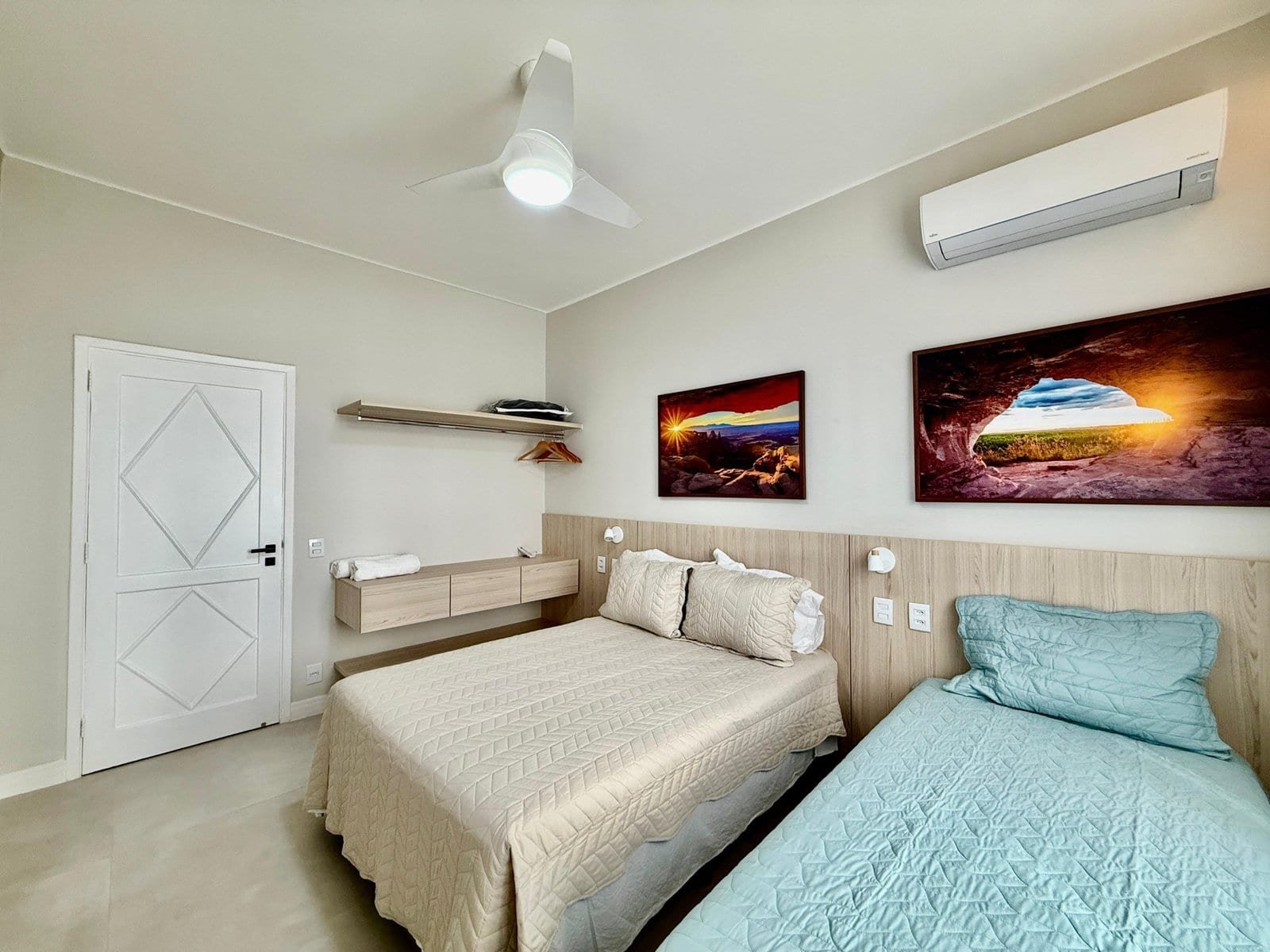 Quarto de hotel com duas camas, decoração moderna, paredes claras, quadros com fotos de paisagens, ar-condicionado e detalhes de iluminação, ideal para descanso confortável.