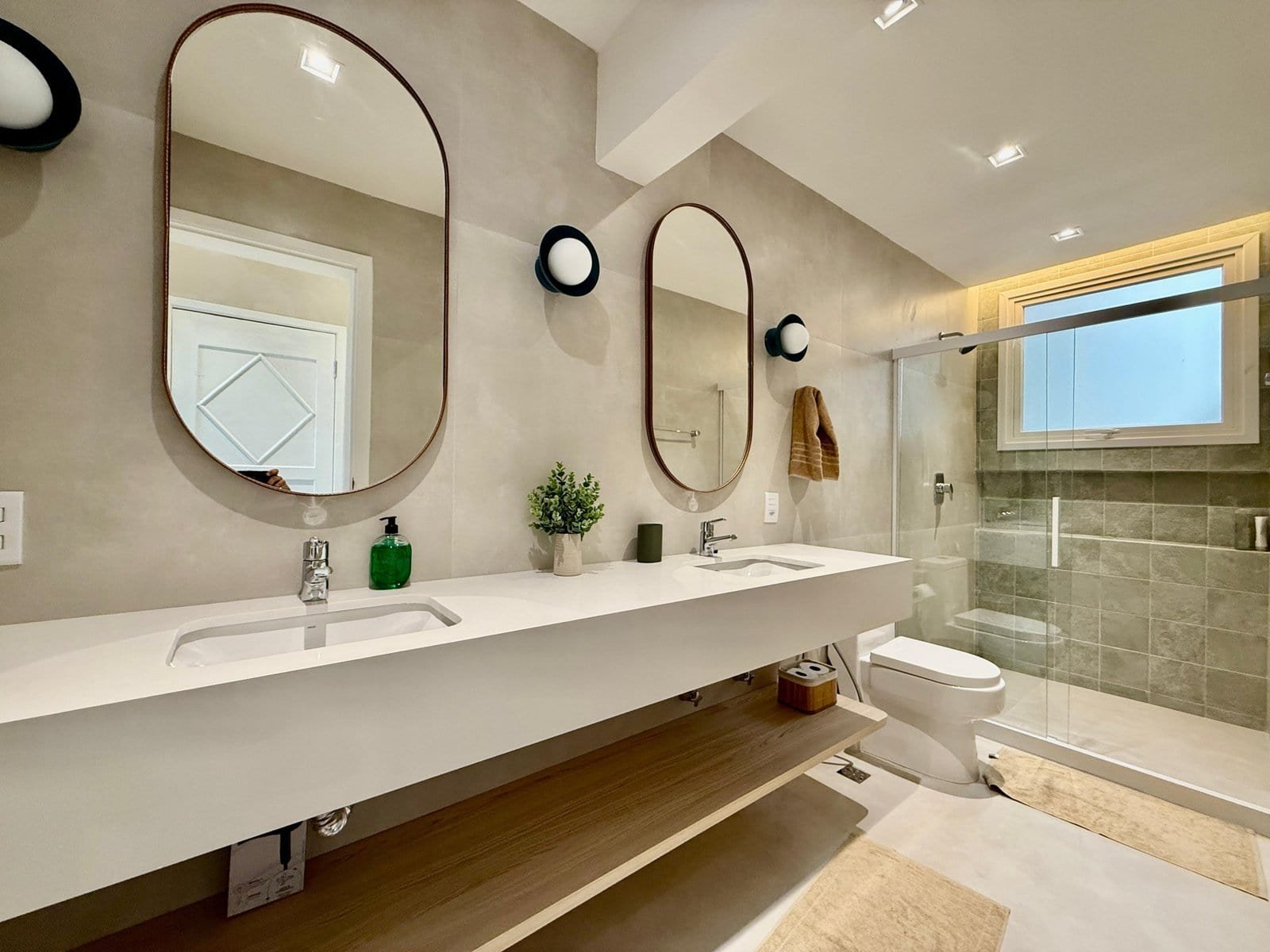 Banheiro moderno com double lavatório, espelhos arredondados, iluminação embutida e ducha com janela de vidro natural. Design elegante e funcional para conforto e praticidade.