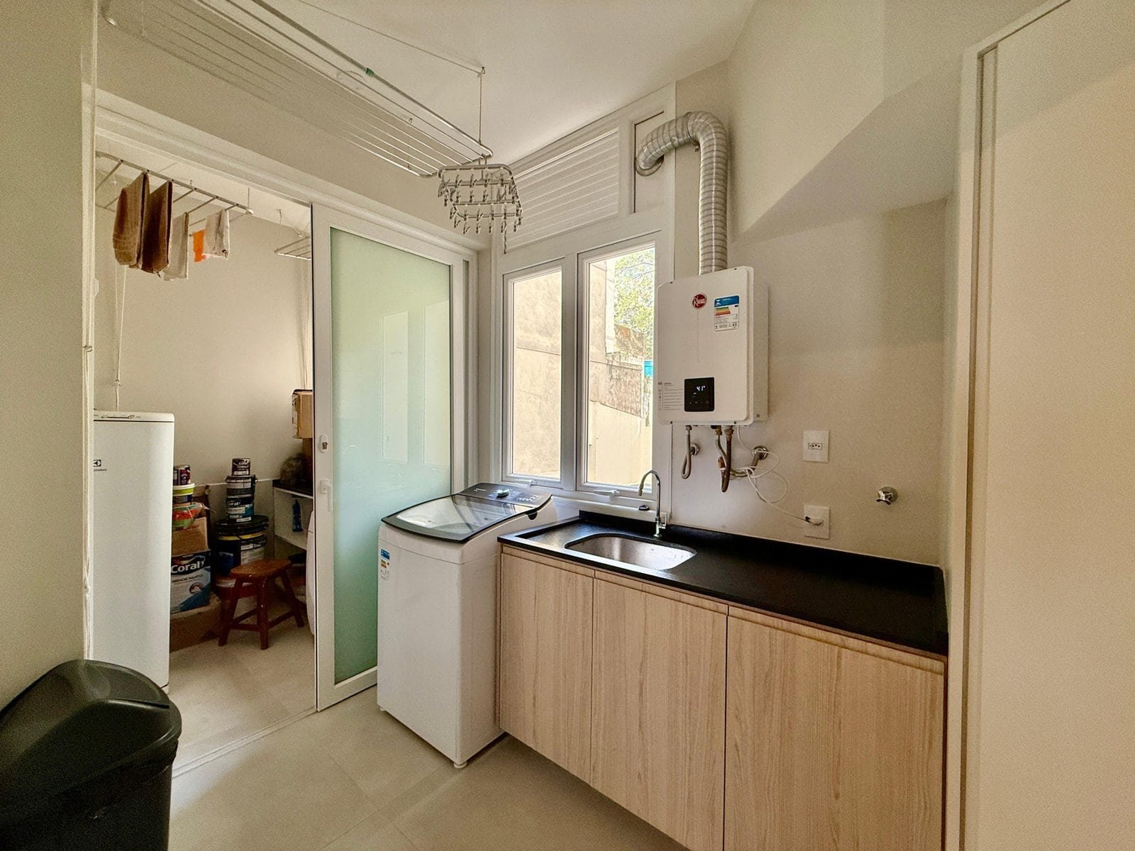 Lavanderia moderna com cuba, máquina de lavar, aquecedor de água e armários de madeira, iluminação natural e janelas grandes, ideal para uso residencial ou comercial, com espaço organizado e funcionalidade.