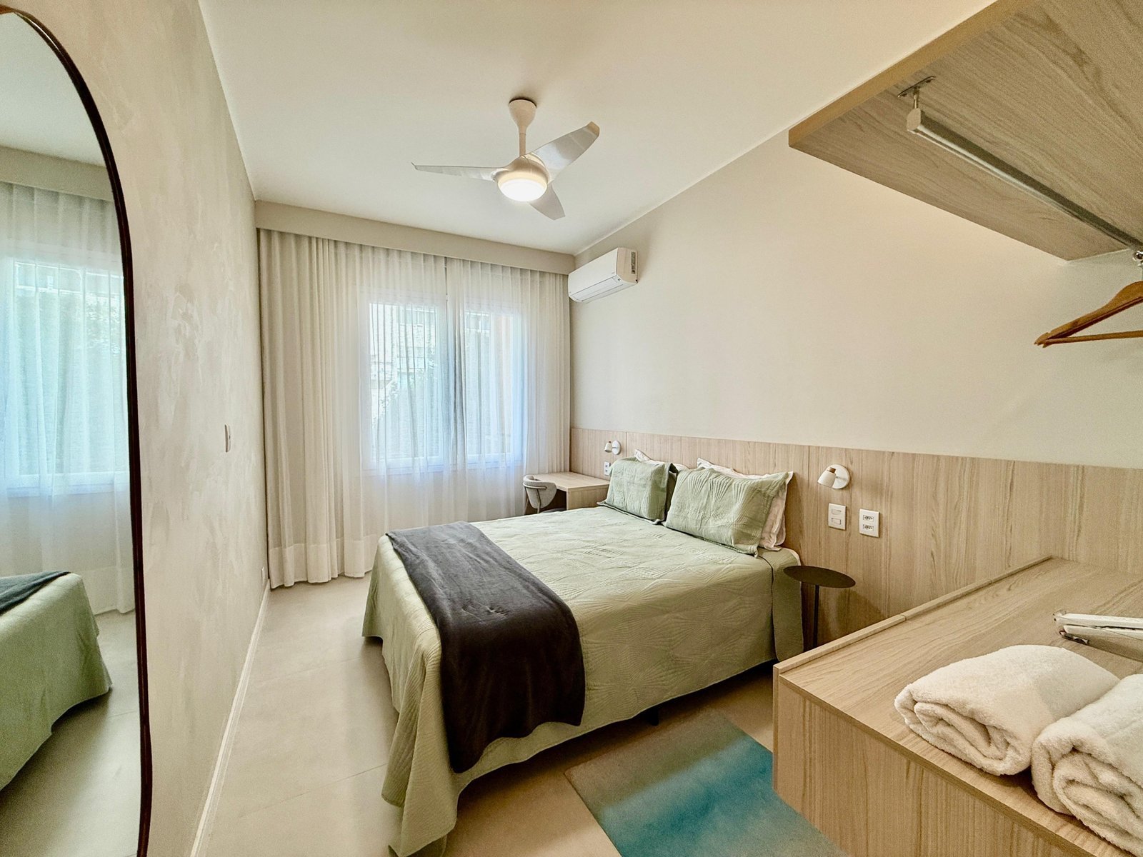 Quarto de hotel com decoração moderna, cama de casal, cortinas de tecido claro, ar-condicionado e iluminação suave, ideal para conforto e relaxamento.