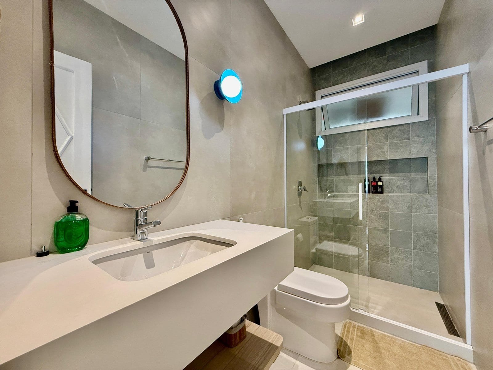 Espelho oval no banheiro moderno com bancada branca, ducha de vidro com revestimento em pedra cinza, iluminação embutida e janela para luz natural.