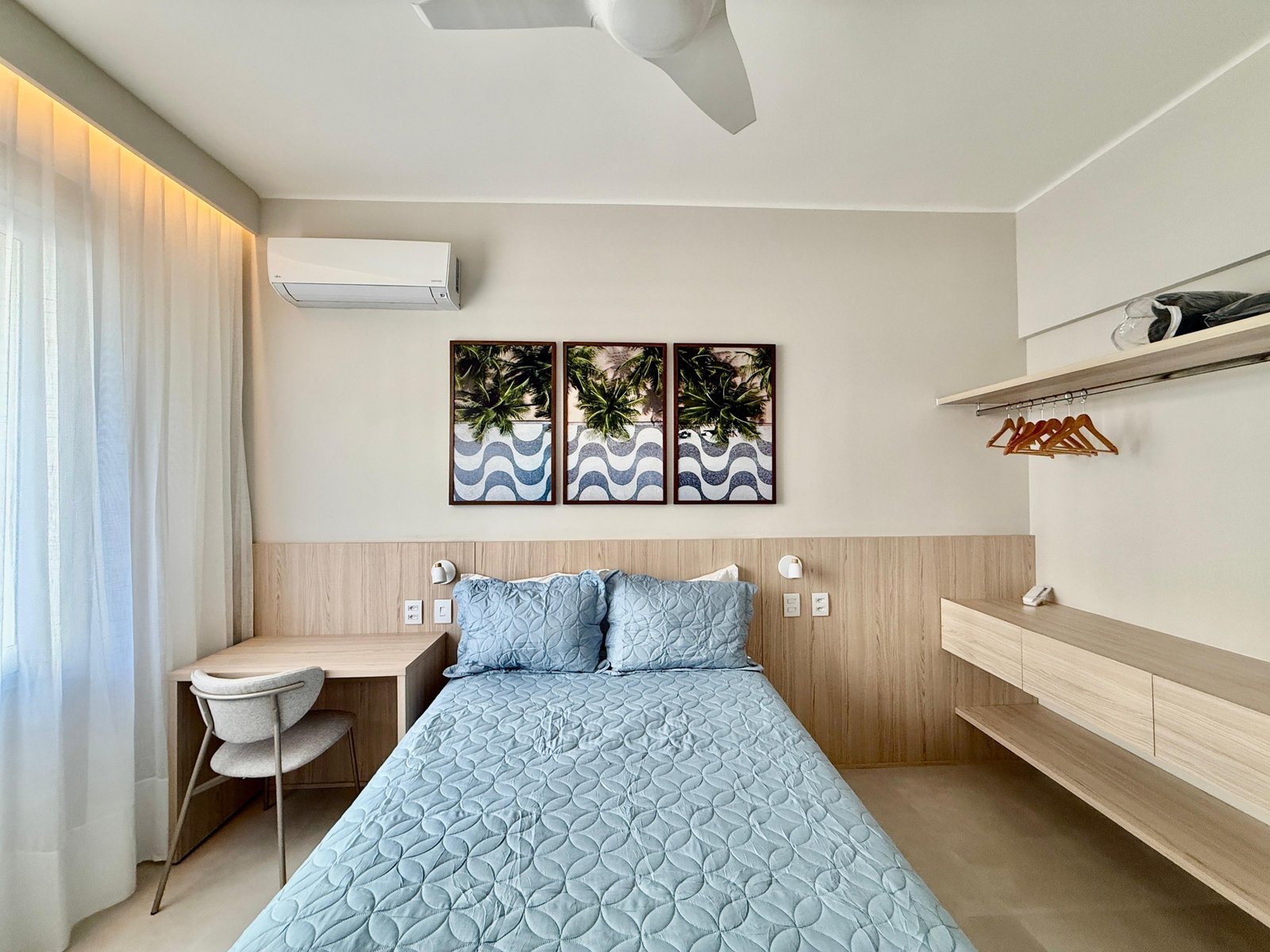 Conjunto de fotos de um quarto de hotel moderno e minimalista, com decoração clara e confortável, incluindo cama de casal com roupa de cama em tom azul, parede com arte moderna, ar-condicionado, bancada de trabalho e armário com cabides.