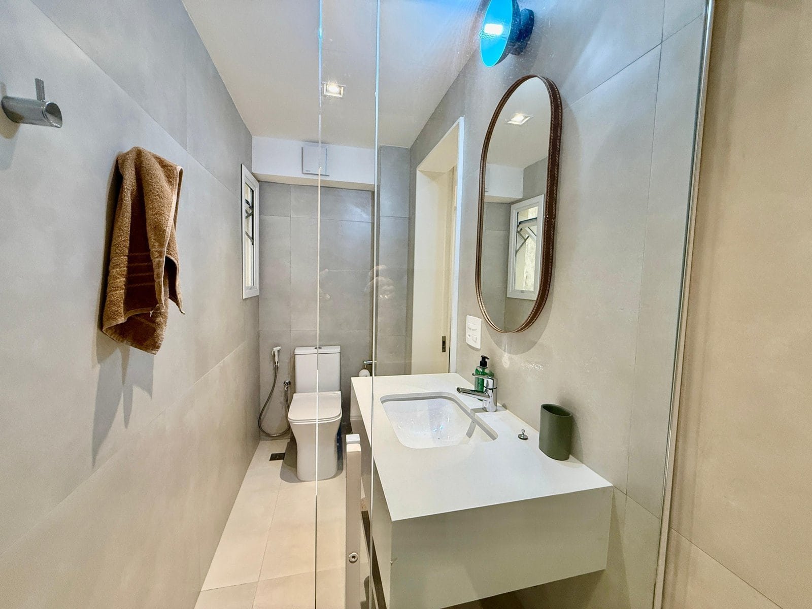 Banheiro moderno com revestimento em tons neutros, espelho oval acima da bancada e toalhas organizadas na parede. Espaço funcional com ducha de vidro e pia com sabonete líquido e porta escova de dente.
