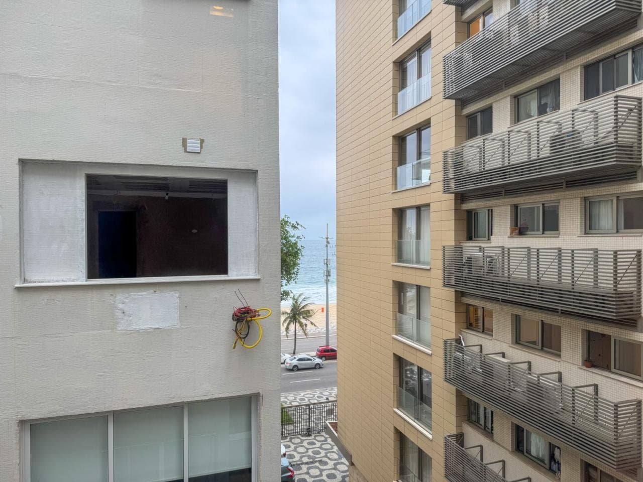 Vídeo de apartamentos residenciais em construção na cor bege, com varandas de metal, vista para a praia e o mar ao fundo, céu nublado, com carros estacionados na rua e árvores ao lado.