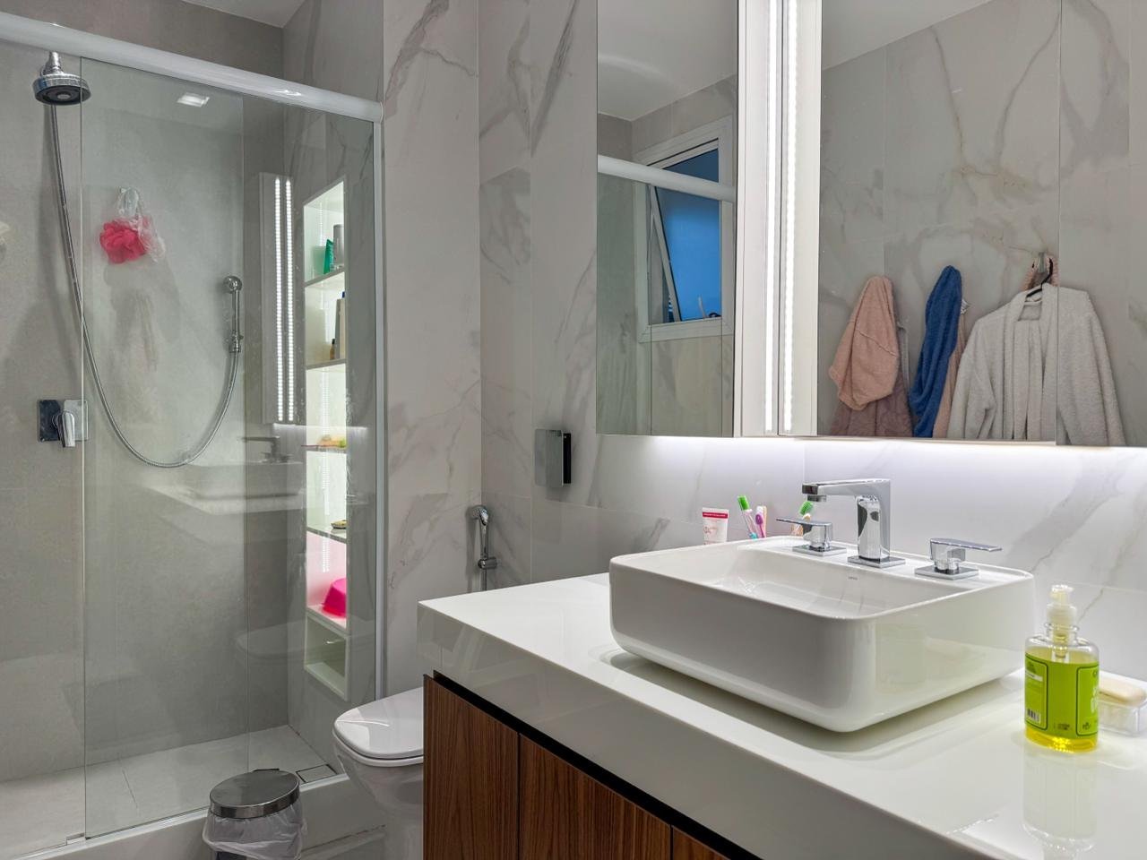 Banheiro moderno com pia retangular, espelho iluminado, detalhes em mármore, box de vidro, toalhas penduradas, espaço bem iluminado, estética clean, ambiente de lavabo de alto padrão.