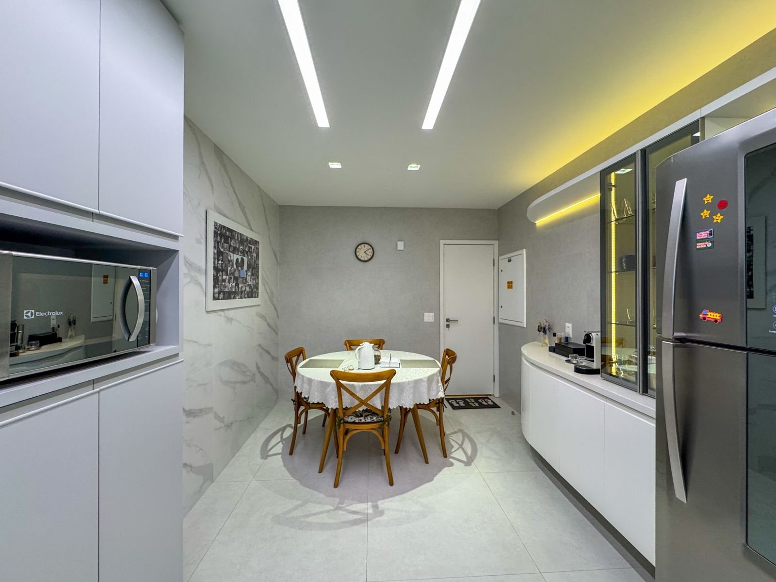 Moderno espaço de copa com móveis brancos, eletrodomésticos inox, mesa redonda com toalha branca, iluminação embutida e decoração minimalista, ideal para ambientes corporativos ou residenciais sofisticados.