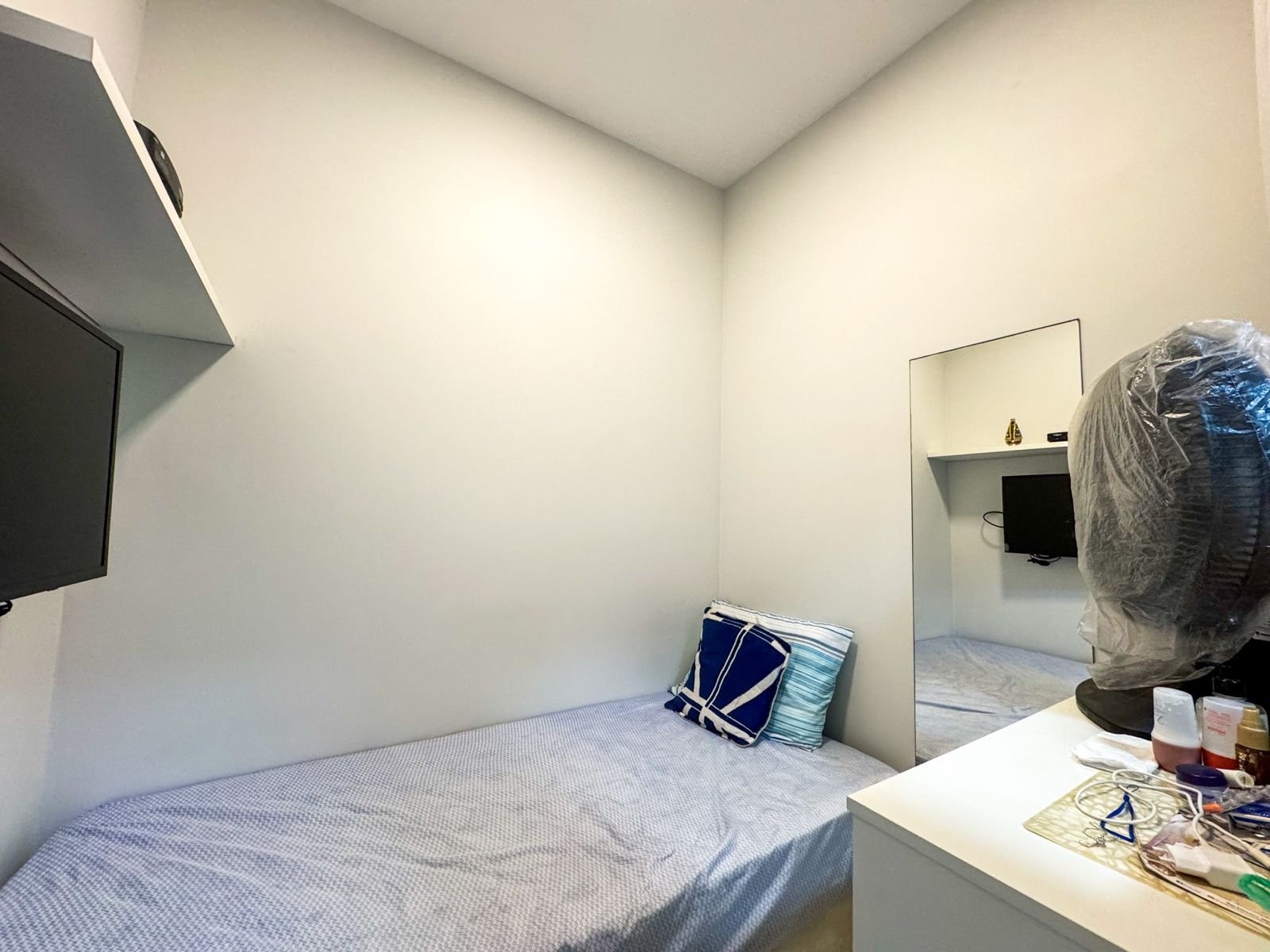 Acontecimento de quarto de hotel simples em ambiente compacto com cama de solteiro, espelho na parede, televisão, objetos pessoais na cômoda e uma camiseta embolada no espelho, decoração minimalista, ambiente confortável para hospedagem.