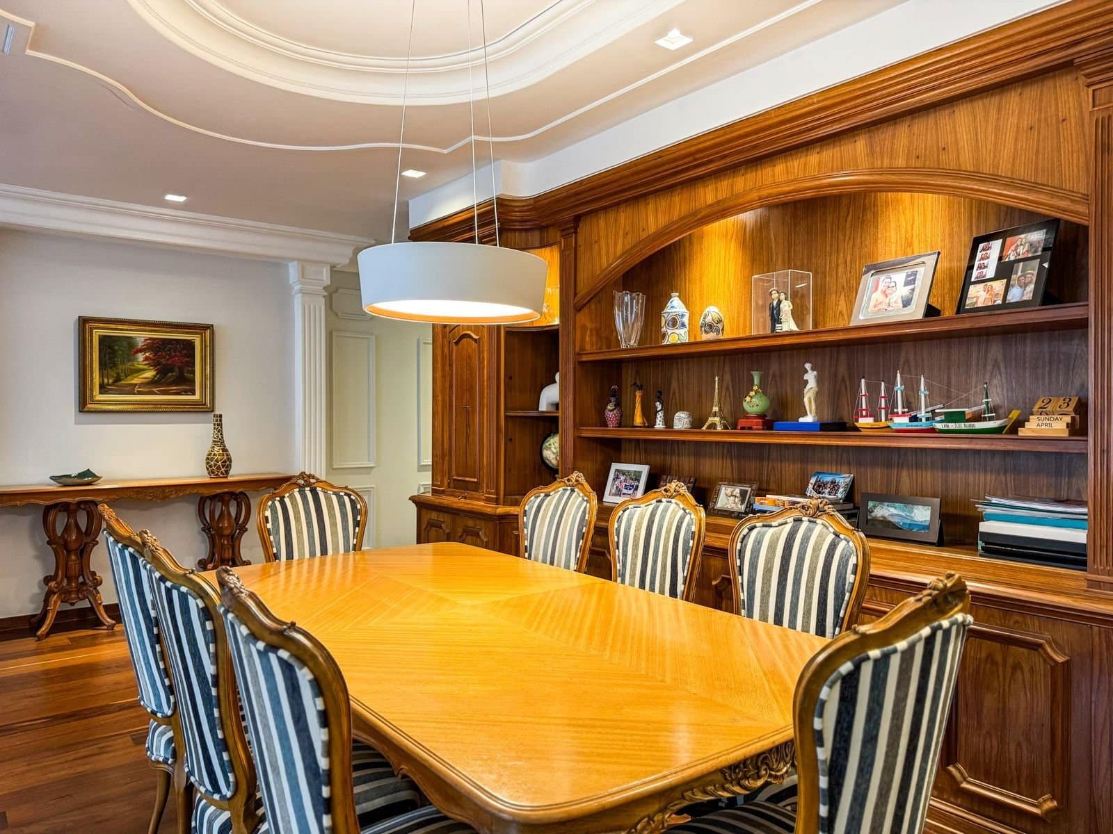 Conjunto de sala de jantar com móveis de madeira, cadeiras estofadas com tecido listrado preto e branco, painel de madeira com fotos e objetos decorativos, iluminação moderna e ambiente sofisticado.